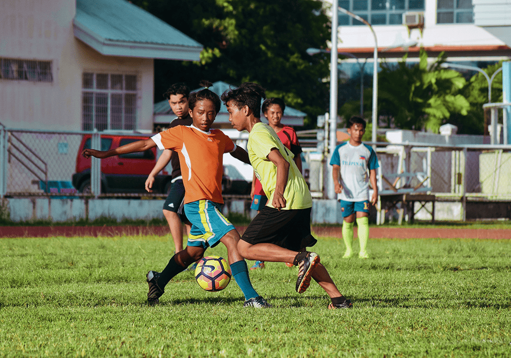 Young players in bright sports attire are playing soccer on a grass field, with a school building in the background.