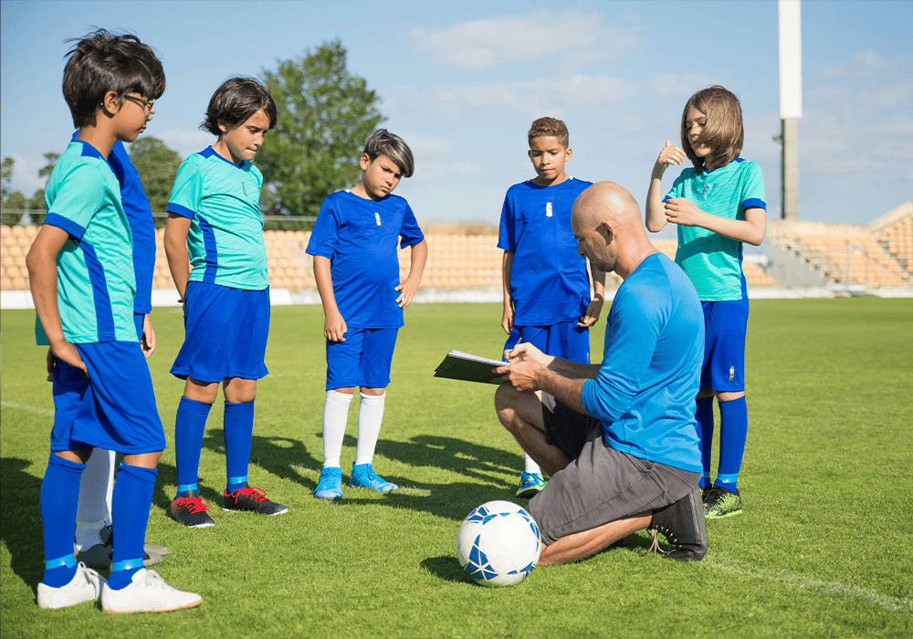 A coach in a blue uniform gives instructions to young soccer players on a sunny field. The players, also in blue uniforms, attentively listen.