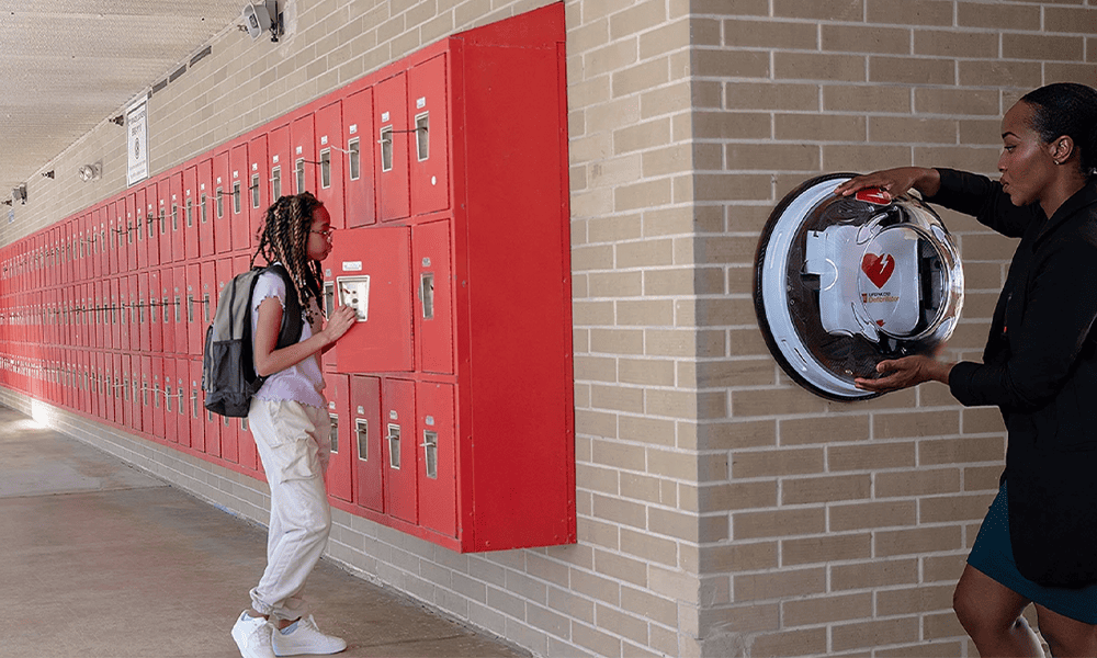 Young girl student stands next to her lockers, on the other wall a woman is attaching an AED to the wall, which is a life-saving tool against sudden cardiac arrest