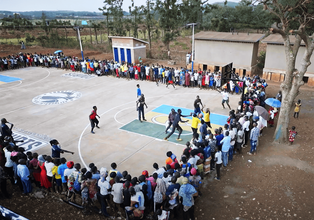 Aerial view of a basketball game in progress on an outdoor court with Shooting Touch and Pat Connaughton Foundation logos, surrounded by a large crowd of spectators.